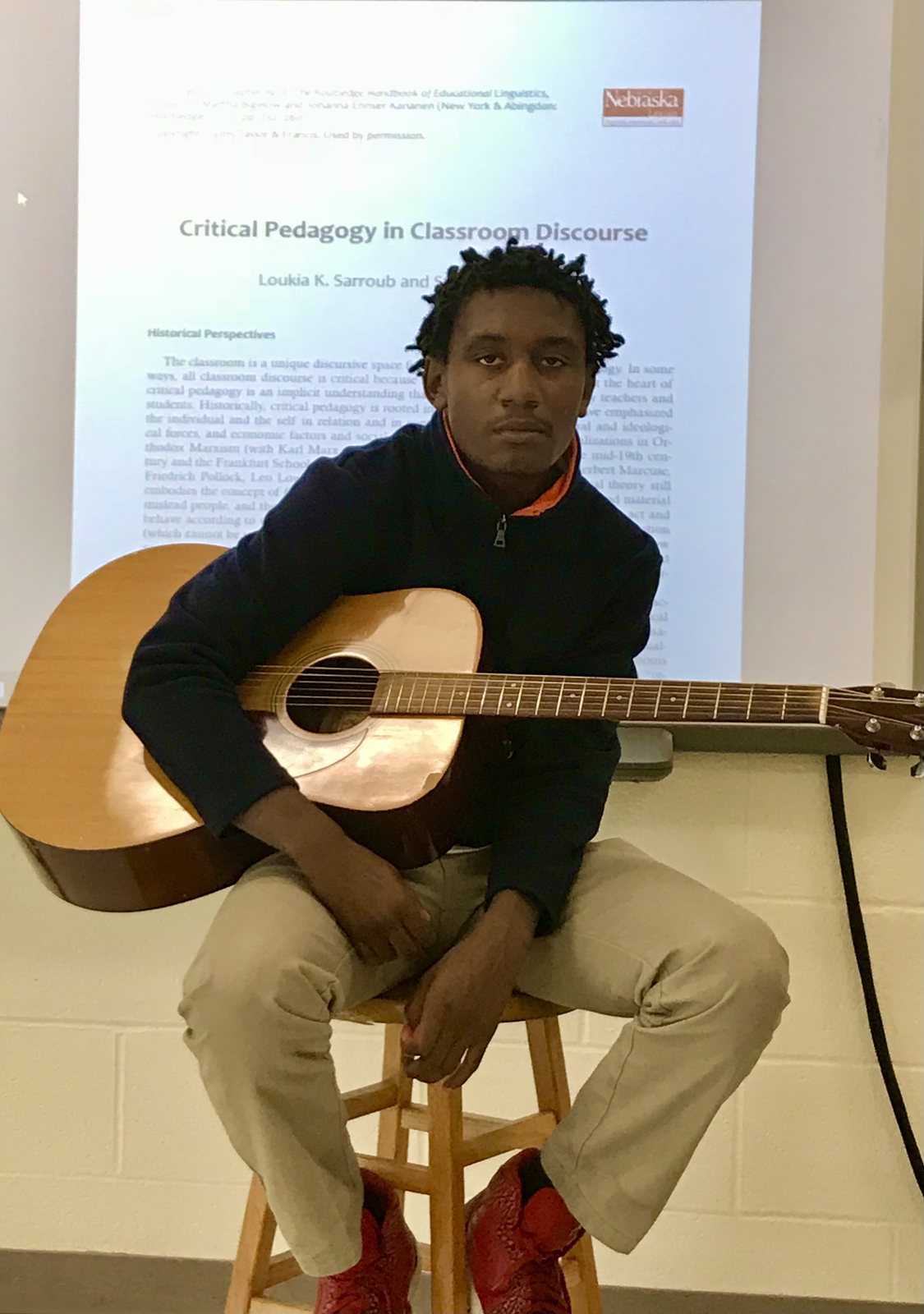 A student seated on a stool holding an acoustic guitar, an article titled Critical Pedagogy in Classroom Discourse projected above his head